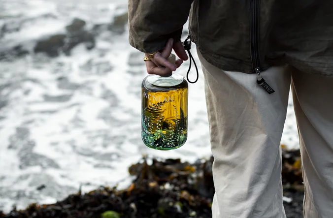 Person holding a decorated Whisky Bottle by the Ocean. Image Credit Nc'nean
