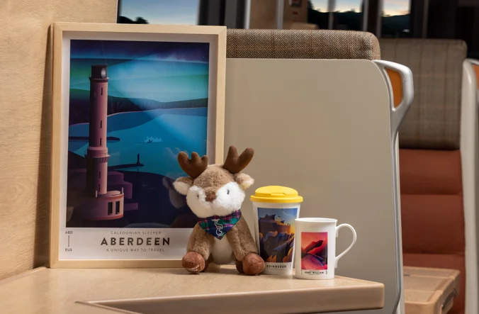 Framed Caledonian Sleeper artwork with stag plushie travel mug and mug in train carriage.