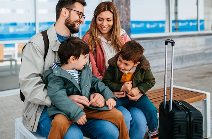 A mother and father with 2 small children sat on a bench with suitcases nearby.